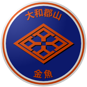 The logo of the Yamatokoriyama Kingyo (Goldfish): a blue circle bordered in white-orange-white, inside of which the orange emblem of Yamatokoriyama, which looks like a complex floor tile, sits between the characters for "Yamatokoriyama" and "Kingyo."